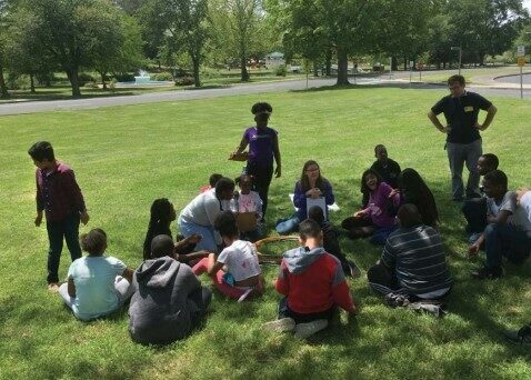 Students gather around their teacher on the lawn of their school grounds.