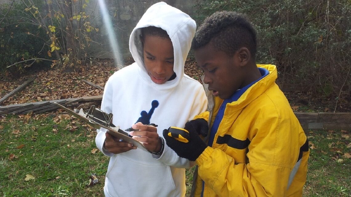 Two young students taking notes on a pad while reading a water quality meter.