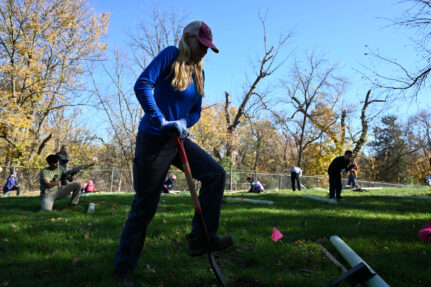 A person uses one leg to push a shovel into the ground in a grass field. In the background, other people are spread out in the field digging and handling tree planting equipment.